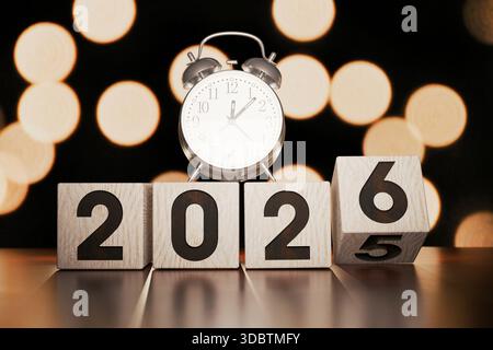 Conto alla rovescia per l'anno nuovo 2026. Una sveglia si trova sui blocchi numerici in legno, simboleggiando l'anno prossimo e la scadenza per gli obiettivi. Festa bokeh Backgrou Foto Stock