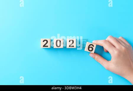 Concetto di pianificazione per il nuovo anno e il futuro. Blocco di legno che cambia a mano da 2025 a 2026 su sfondo blu, simboleggia un nuovo inizio, obiettivi aziendali e visione Foto Stock