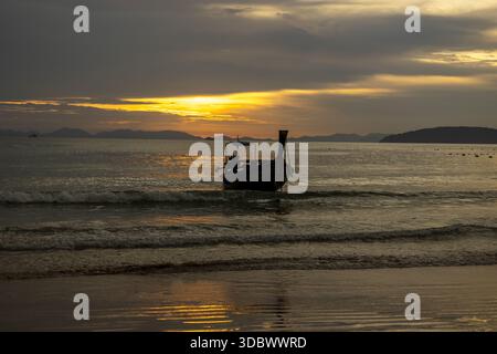 La vista della silhouette di una barca a coda lunga si trova serenamente sull'acqua mentre il sole tramonta, proiettando un bagliore dorato sulle onde ondulate, Ao Nang, Krabi e Thaila Foto Stock