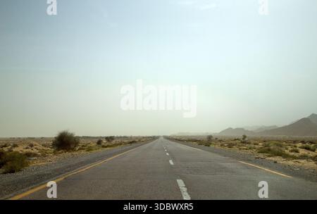 Vista di una lunga e dritta autostrada che attraversa il paesaggio arido sotto un cielo nebbioso, con vegetazione sparsa e montagne lontane, Makran Coastal Hig Foto Stock