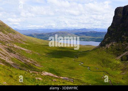Rigogliosa e lussureggiante valle con tortuosi scricchioli al centro e ripide scogliere rocciose e erbose su ogni lato alla luce del sole, lago e montagne collinari nel Foto Stock