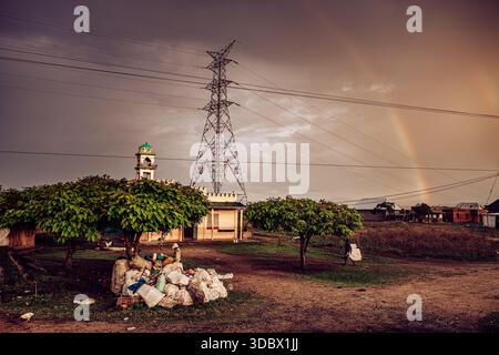 La vista di una moschea si erge accanto a un pilone elettrico sotto un cielo drammatico con un arco arcobaleno in testa, spazzatura sparsi in primo piano, Kampala, centesimo Foto Stock