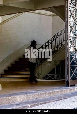 Un uomo che indossa un cappello e un abbigliamento casual si trova vicino a una scala, con un'altra persona seduta su un gradino in primo piano. Foto Stock