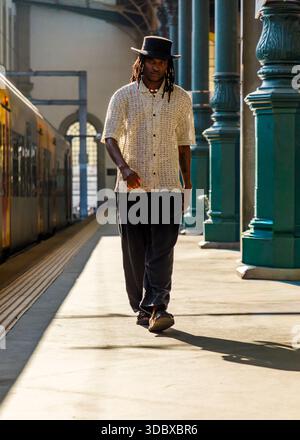 Un uomo che indossa un cappello e una camicia con bottoni cammina su una piattaforma del treno. Foto Stock