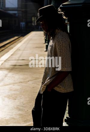 Una persona che indossa un cappello e un abbigliamento casual si trova su una piattaforma del treno, appoggiata a una struttura scura. Foto Stock