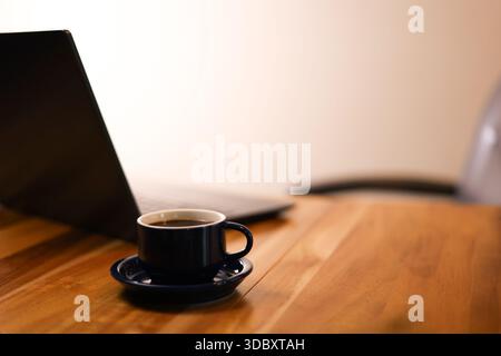 Tazza da caffè messa a fuoco con laptop e sedia sfocati sullo sfondo Foto Stock
