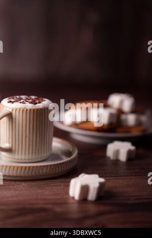 ambiente accogliente di cappuccino in tazza a costine con schiuma a motivi geometrici, accompagnato da forme decorative di zucchero e biscotti su un tavolo in legno, contro il marrone caldo Foto Stock