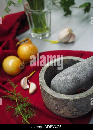 Mortaio e pestello in pietra con grani di pepe, aglio e cipolla su sfondo in tessuto rosso Foto Stock