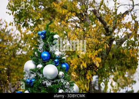 Un albero di Natale decorato con ornamenti blu e argento sorge all'aperto accanto al fogliame autunnale e alle piccole luci Foto Stock