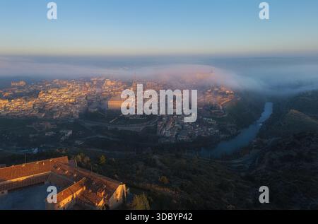 L'intero skyline di Toledo si illumina sotto la calda luce dell'alba e la nebbia. Il Alcázar e la cattedrale emergono dalla nebbia in tonalità dorate. Foto Stock