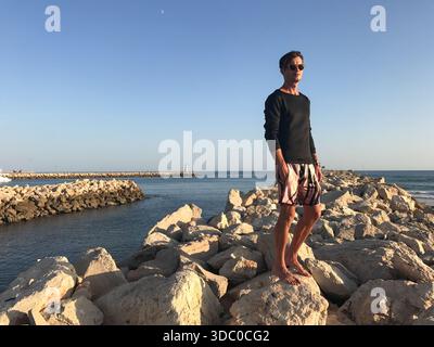 Bel modello maschile si erge con sicurezza su rocce naturali formando uno stretto sentiero lungo un fiume che scorre verso il mare. Indossa un elegante outfit casual Foto Stock
