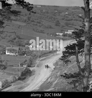 Bohorodychne, regione di Donetsk, regione di Sloviansk, RSS Ucraina, URSS, 1960 anni Una fotografia archivistica di paesaggi ad alto angolo di una valle rurale. Una tortuosa strada sterrata attraversa il villaggio, con un carro trainato da cavalli che lo percorre. Le tradizionali case dipinte di bianco con giardini recintati sono sparse attraverso le dolci colline. Un tronco di pino in primo piano incornicia la scena. L'immagine cattura l'atmosfera idilliaca, i trasporti tradizionali e la vita quotidiana della tranquilla campagna del Donbas. Foto Stock
