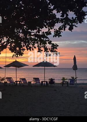 Le sedie a sdraio fiancheggiano la spiaggia sabbiosa di Baan Krood mentre il sole tramonta all'orizzonte. Gli ombrelli offrono ombra mentre il cielo si riempie di colori vivaci Foto Stock
