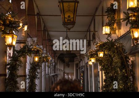 Rovigo, italia 18 dicembre 2025: Lanterne illuminate e ghirlande verdi che decorano un vecchio passaggio ad arco, creando un ambiente festoso, elegante e tranquillo Foto Stock