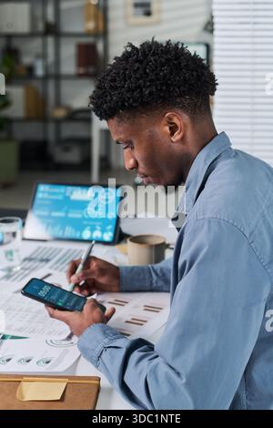 Giovane uomo nero adulto che lavora alla scrivania analizzando i dati finanziari su smartphone e tablet digitale, tenendo la penna e rivedendo grafici stampati, concentrandosi sul multitasking negli uffici moderni Foto Stock