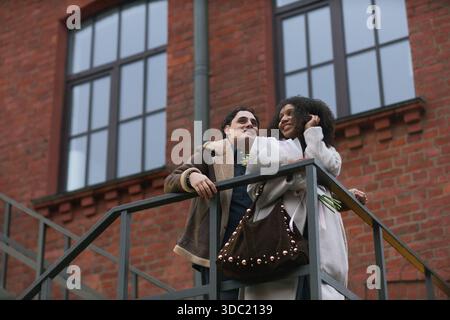 Giovane uomo caucasico adulto e giovane donna nera adulta in piedi insieme su una scala all'aperto, sorridendo e tenendo fiori, godendo momenti romantici in un ambiente urbano, un contesto di incontri Foto Stock