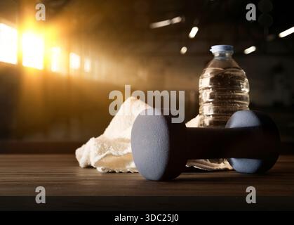 Concetto di sport, allenamento e benessere con due manubri in neoprene blu su una panca di legno con una bottiglia d'acqua e un asciugamano bianco dietro e palestra scura bac Foto Stock