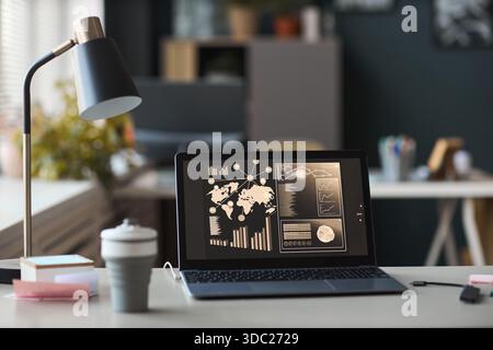 Spazio di lavoro moderno con laptop aperto che visualizza dashboard di analisi aziendale con mappa del mondo, grafici e grafici sullo schermo, lampada da scrivania e forniture per ufficio che circondano il dispositivo Foto Stock