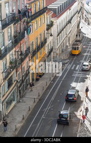 Vista dall'alto dell'iconico tram giallo 28 che attraversa una stradina stretta di Lisbona, Portogallo, circondata da edifici colorati e automobili. Foto Stock