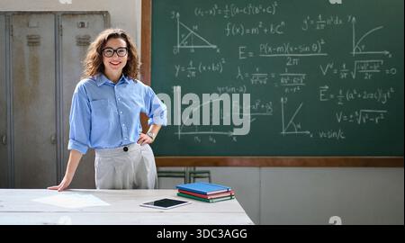 Una giovane insegnante di matematica a scuola sta alla scrivania di fronte alla lavagna in classe. Foto Stock