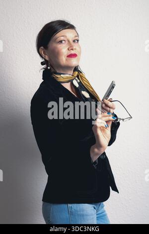 Una donna con una giacca nera e degli occhiali ha un cellulare. Indossa una sciarpa gialla e ha lo smalto blu Foto Stock