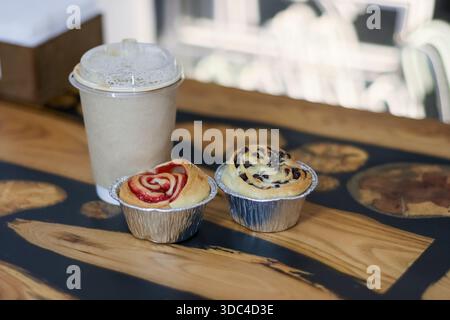 Muffin alla rosa alla fragola e muffin con gocce di cioccolato in tazze di alluminio con caffè ghiacciato su un tavolo in legno di resina artistica Foto Stock