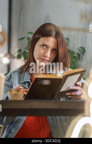 Primo piano di una giovane donna con giacca in denim profondamente impregnata di un libro da lettura con caffè sul tavolo Foto Stock