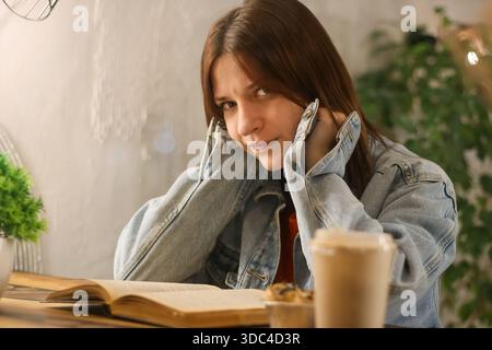 Giovane donna premurosa in giacca di denim che riposa stancante al tavolo del caffè con caffè da asporto e pasticcini Foto Stock