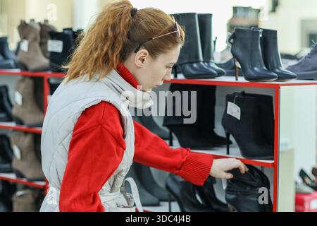 Vista laterale del cliente che sta cercando uno stivaletto nero sul ripiano rosso in un negozio di scarpe affollato. Foto Stock