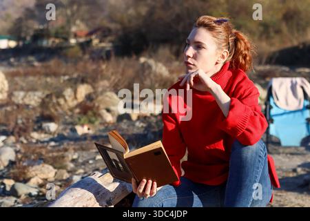 Profilo di una giovane donna in rosso, mano sul mento, avvolto in libro su un ambiente roccioso all'aperto Foto Stock