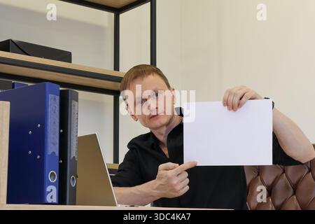 Un giovane uomo adulto in camicia nera alla scrivania dell'ufficio con laptop e cartelle, alzando carta bianca bianca bianca e puntando l'indice verso di essa. Foto Stock