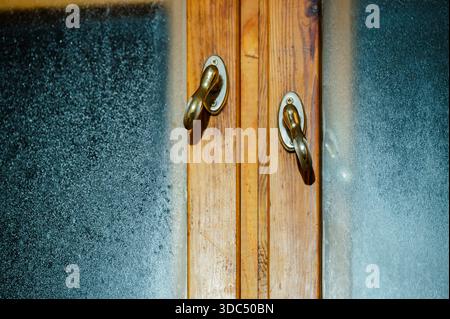 Wooden window frame showing two brass handles and frosted glass covered in condensation and ice crystals, symbolizing cold winter weather and home com Foto Stock