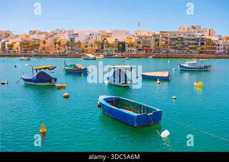 Il porto di Marsaxlokk, Malta, è affollato di colorate barche da pesca luzzu in acque turchesi. Pittoreschi edifici sul lungomare completano la soleggiata sce Mediterranea Foto Stock
