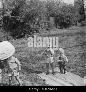Regione di Donetsk, RSS Ucraina, URSS, metà degli anni '1970 Foto d'archivio in bianco e nero di bambini che giocano in un giardino rurale. Visto da dietro le spalle di un adulto, un ragazzo mangia uno spuntino mentre è in piedi su una coperta accanto a una bambina con un grande arco di capelli. Un altro bambino cammina nelle vicinanze. Una vivace scena della vita familiare sovietica e dei fratelli che interagiscono all'aperto in estate. Autentica fotografia cinematografica d'epoca. Foto Stock