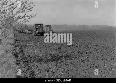 Regione di Donetsk, RSS Ucraina, URSS, metà degli anni '1970 Foto d'archivio industriale in bianco e nero di coltivazione di terreni pesanti. Due trattori caterpillar sovietici (tipo T-74 o DT-75) tirano erpici a disco in un vasto campo agricolo collettivo. Le macchine si spostano in tandem, accumulando polvere e girando grandi strati di terra. I giovani alberi sono in primo piano. Un potente scenario di aratura primaverile e meccanizzazione agricola nella steppa. Autentica fotografia cinematografica d'epoca. Foto Stock