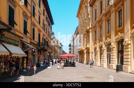 Pisa, Italia - 26 giugno 2025: Gli edifici illuminati dal sole fiancheggiano corso Italia mentre la gente passeggia accanto ai negozi e si riunisce sulla terrazza centrale con il suo ombrello rosso Foto Stock