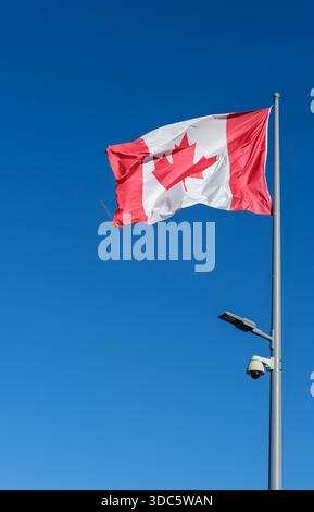 Vista esterna ad angolo basso della bandiera canadese rossa e bianca su asta metallica, struttura in tessuto visibile, luce stradale montata e telecamera di sicurezza a cupola. Foto Stock