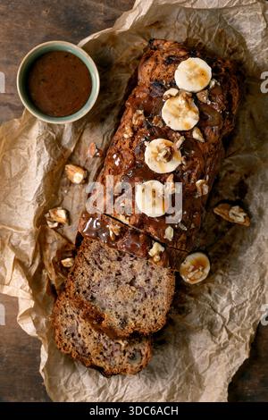 Pane alla banana fatto in casa a fette con noci, condito con caramello e condito con banana fresca. Su carta pergamena increspata con una ciotola di salsa Foto Stock