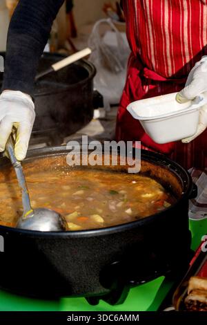 Festival europeo del cibo di strada. Fornitore che indossa guanti usa e getta che serve la zuppa tradizionale di patate da una pentola grande alla ciotola da asporto. Autentica cucina casalinga Foto Stock
