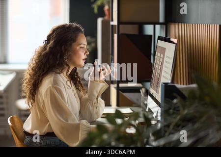 Giovane donna caucasica adulta seduta alla scrivania con assistente vocale sullo smartphone mentre lavora al computer in un ufficio moderno, espressione mirata, capelli ricci, vista laterale Foto Stock