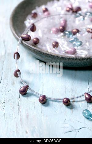 Creazione di gioielli fatti a mano. Creazione di collana con perline in vetro e componenti in una ciotola in ceramica su sfondo di legno chiaro. Concetto di hobby fai da te. Chiudi Foto Stock