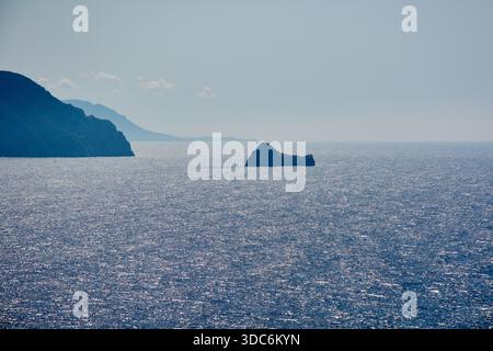Una vista mozzafiato della scintillante superficie del Mar Ionio con un piccolo isolotto roccioso e profilo costiero a Corfù, Grecia, raffigurante un tranquillo summ Foto Stock