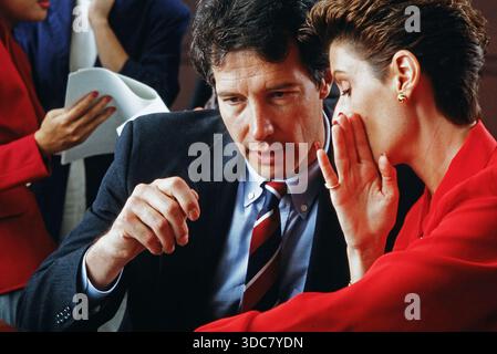 Due dirigenti sono impegnati in una conversazione al tavolo di una riunione mentre i colleghi lavorano nelle vicinanze di un ufficio. E' giorno e i documenti sono sparsi Foto Stock
