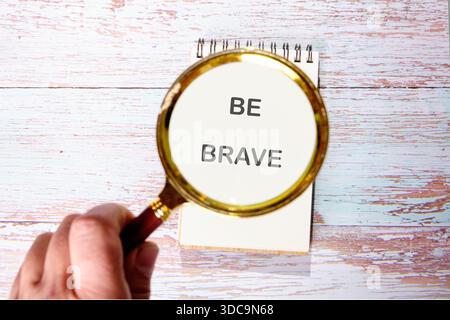 Una mano regge una lente d'ingrandimento su un blocco note appoggiato su una superficie in legno. Il blocco note ha la frase BE BRAVE scritta sulla sua pagina superiore, incoraggiante b Foto Stock