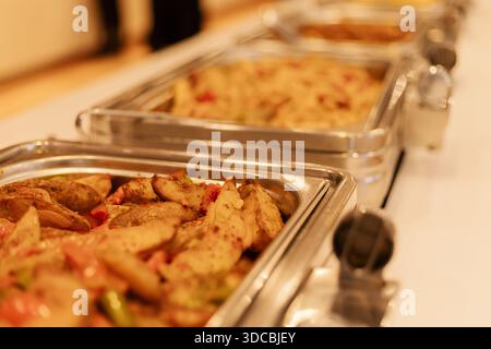 Diversi vassoi metallici pieni di diverse opzioni di cibo sono disposti su un tavolo durante una riunione in una sala conferenze. I vassoi sono organizzati e pronti per Foto Stock