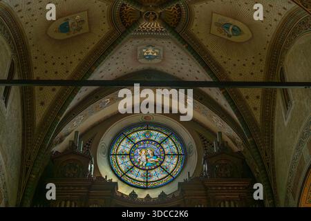 Una vetrata circolare domina l'interno di una chiesa fioca, incorniciata da archi gotici e ricchi lavori in legno. Intricati affreschi sul soffitto e dettagli decorati Foto Stock