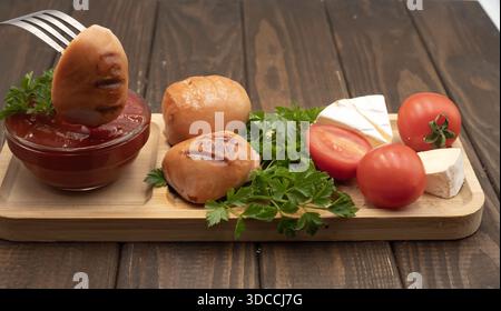 Salsicce grigliate con pomodori, formaggio e ketchup su un piatto di legno, cibo rustico fatto in casa Foto Stock