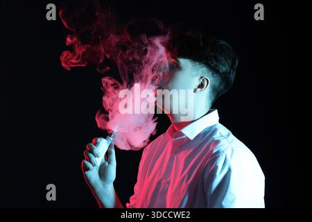 Bel giovane che fuma sigaretta elettronica su sfondo scuro Foto Stock