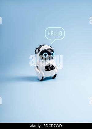 Simpatico robot giocattolo futuristico con occhi brillanti e bolla che dice "HELLO" isolato su sfondo blu, che rappresenta l'assistente ai, la tecnologia chatbot Foto Stock