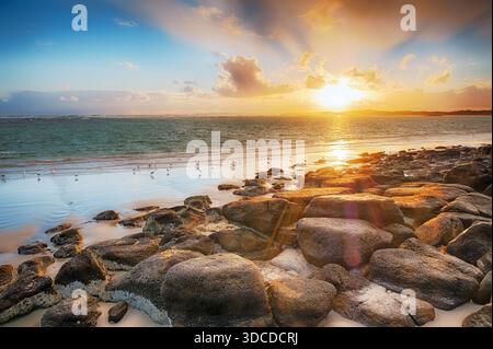 Un tranquillo tramonto sulla spiaggia con rocce in primo piano, dolci onde e uccelli sparsi lungo la riva Foto Stock